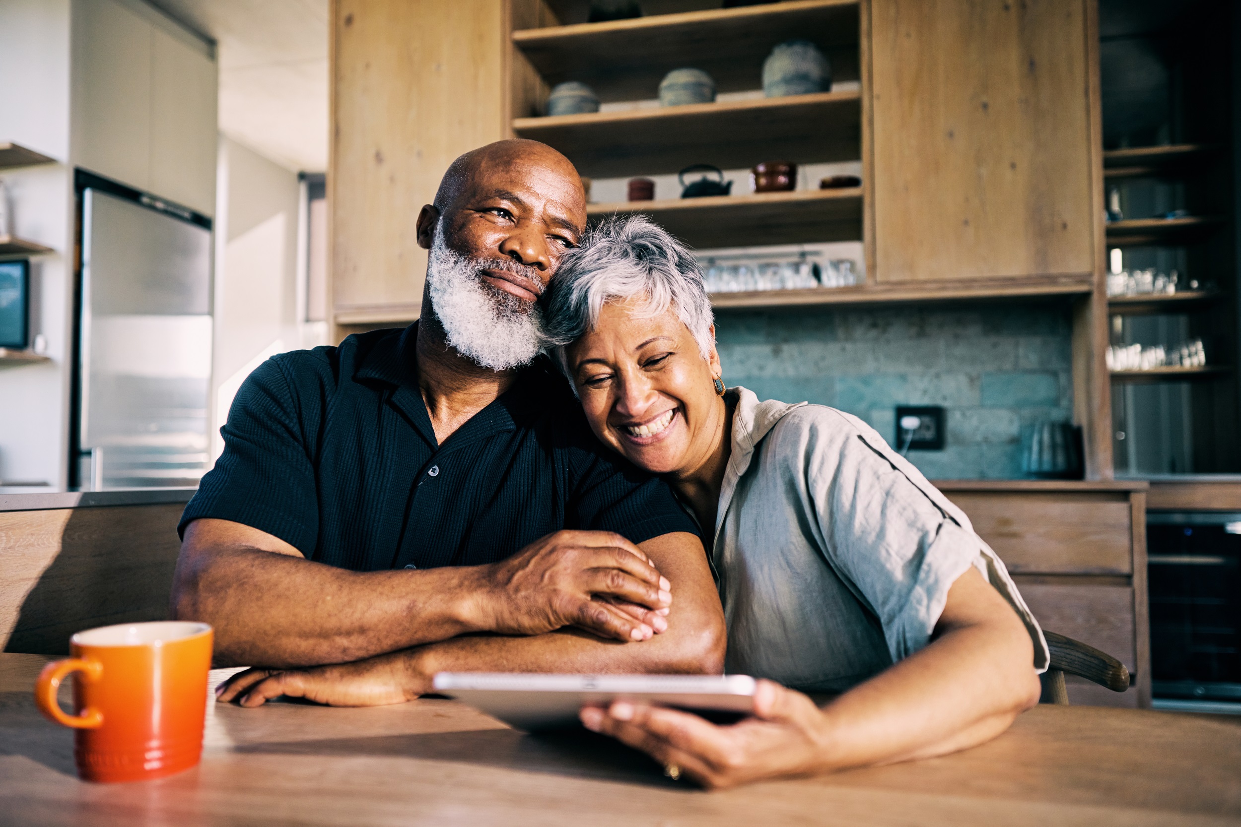 An older couple smiling at a tablet in a cozy kitchen, with a cup of coffee in front of them.