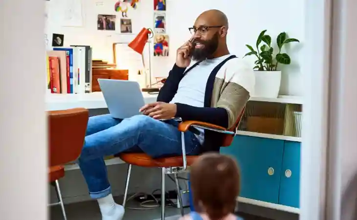 A man in casual dress leans back in the chair of a home office while speaking on a cellular phone with a laptop in his lap.