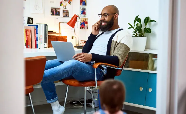 A man in casual dress leans back in the chair of a home office while speaking on a cellular phone with a laptop in his lap.