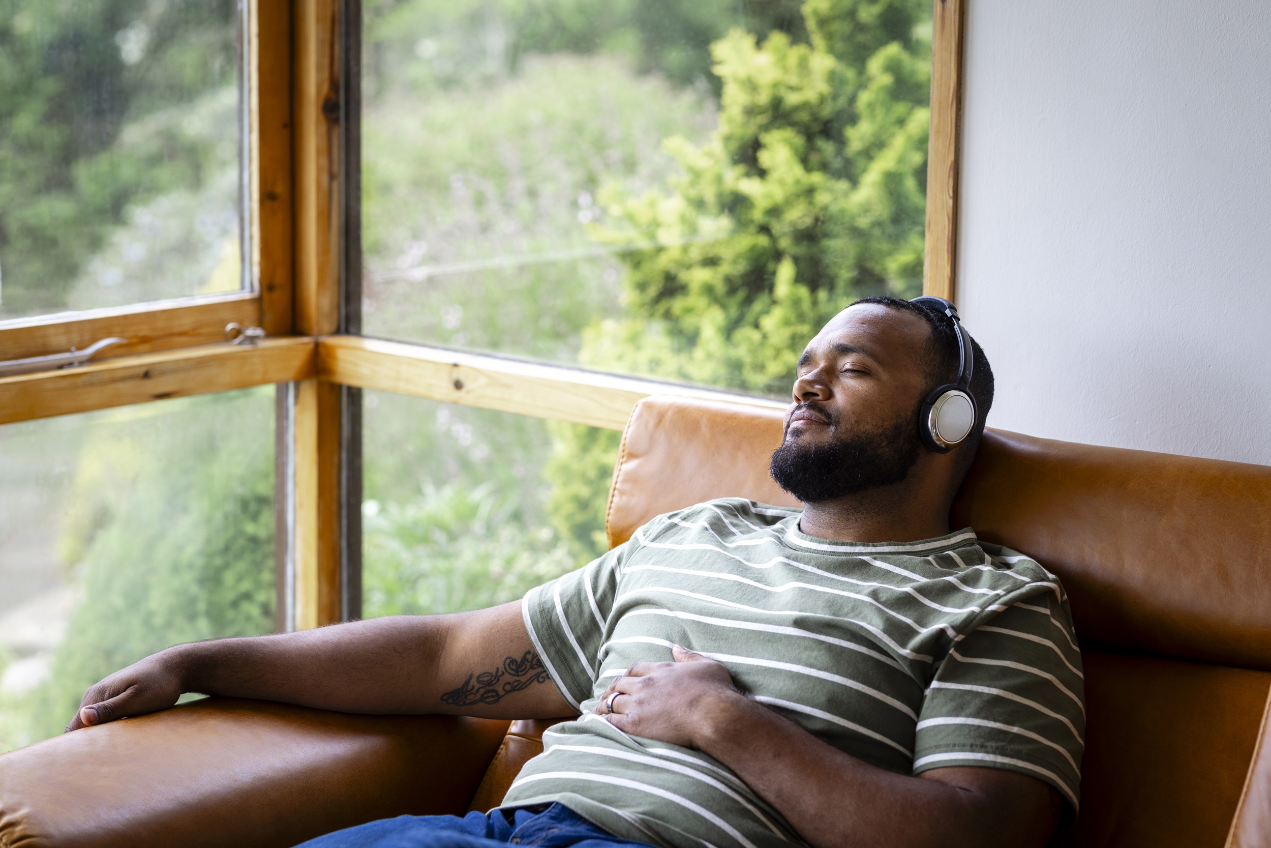 A man relaxing on a leather sofa, wearing headphones with his eyes closed, next to a large glass window.