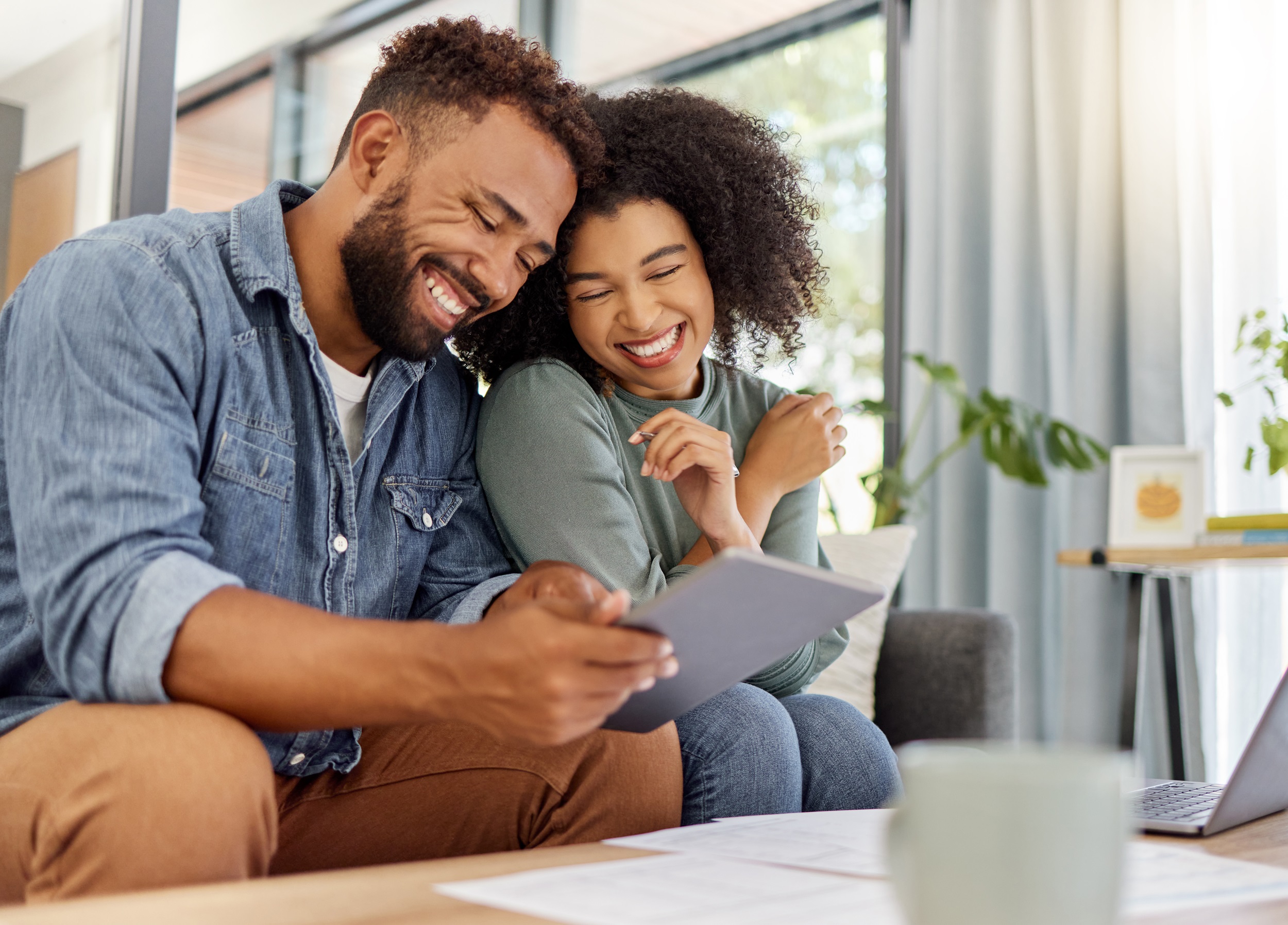 A man and woman smiling at a tablet on a couch, with a laptop and mug on the table in front of them.