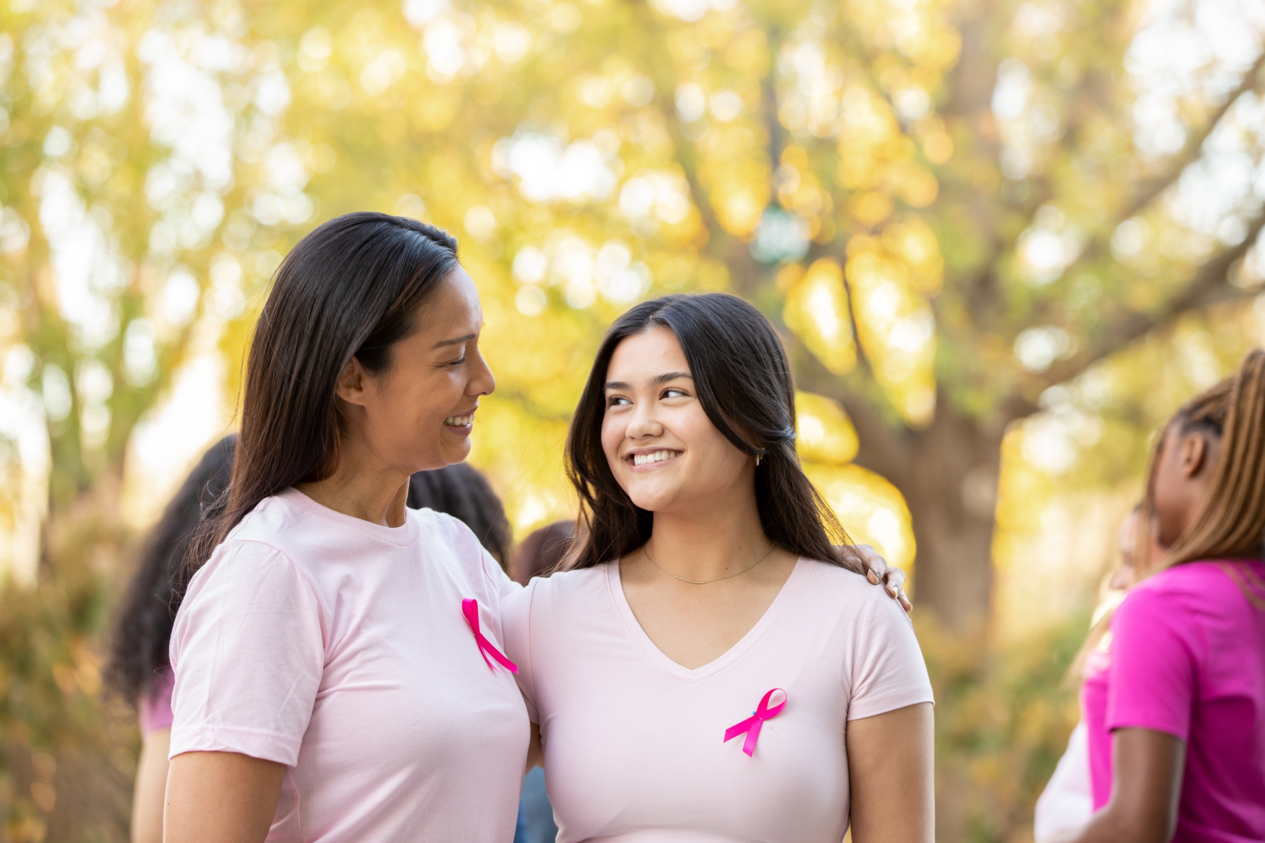 Two women in pink shirts with breast cancer ribbons, standing outside with others in the background.