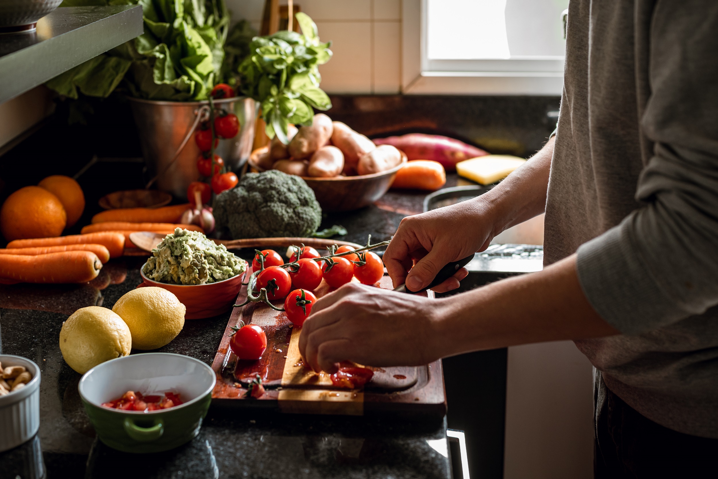A person slicing tomatoes on a cutting board in the kitchen, with various vegetables scattered across the countertop.