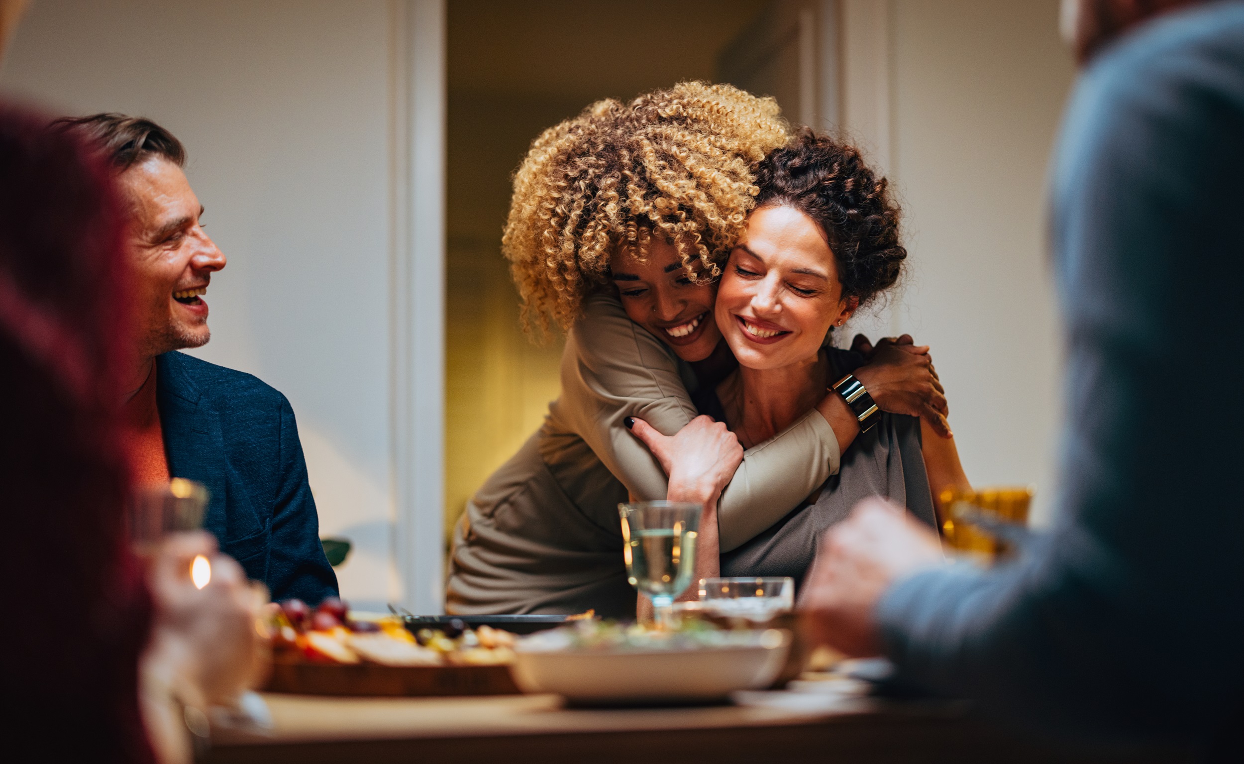 A group of people smiling around a dinner table, with one woman hugging another in celebration.
